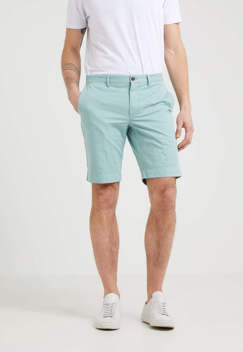 Homme portant un short bleu clair, des baskets blanches et un t-shirt blanc, debout avec une main dans la poche devant un fond uni.