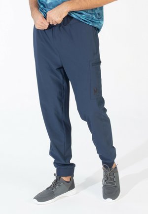 Homme portant un pantalon de jogging bleu marine avec les chevilles retroussées et des baskets grises, ajustant la taille sur un fond blanc.