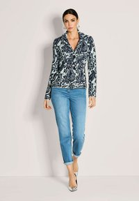 Frau trägt eine gemusterte marineblaue und weiße Jacke mit Knöpfen, hellblaue, hochgekrempelte Jeans und silberne High Heels, steht vor einem schlichten Hintergrund.
