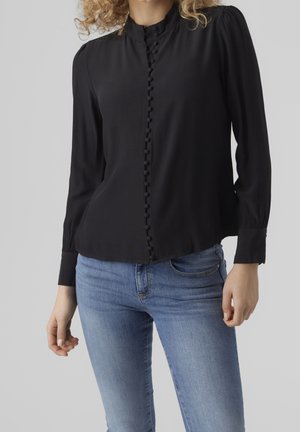 Kobieta ubrana w czarną bluzkę z długim rękawem z obszytymi tkaniną guzikami i niebieskie dżinsy na jednolitym szarym tle.