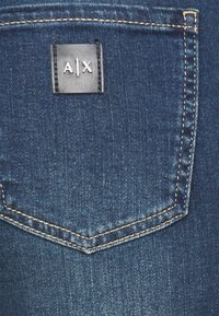 Bolsillo trasero de unos vaqueros de mezclilla azul con costuras beige y una etiqueta cuadrada negra con el logo blanco "A|X".