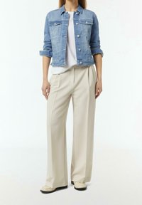 Persona che indossa una giacca di jeans chiara sopra una camicia bianca, abbinata a pantaloni beige a gamba larga e scarpe chiare, in piedi su uno sfondo neutro.