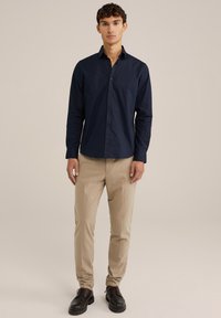 Mornarska srajca z dolgimi rokavi, iz bombažnega materiala, oprijet kroj, v kombinaciji s svetlim bež krojenimi hlačami in črnimi usnjenimi čevlji, stoji ob nevtralni ozadju.