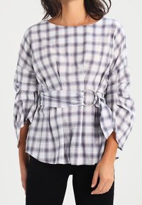 Blusa a quadri bianca e navy con maniche a campana, vita strizzata e dettaglio in anello argentato. Tessuto morbido con una vestibilità rilassata.