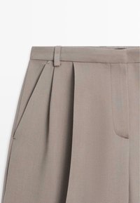 Beige tailored trousers with front pleats, side pockets, and a smooth texture. Features a belt loop waistband and subtle stitching details.