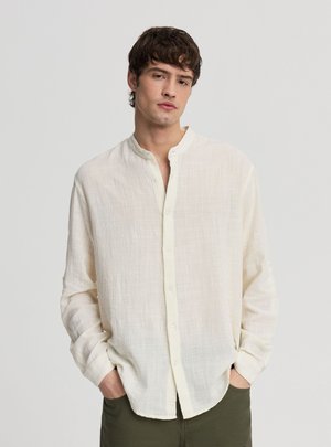 Młody mężczyzna z ciemnymi włosami, ubrany w białą teksturowaną koszulę na guziki i zielone spodnie, stoi z rękami w kieszeniach na jednolitym tle.