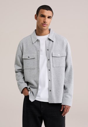 Jeune homme portant une veste boutonnée gris clair sur une chemise blanche, une main dans la poche, sur un fond beige uni.