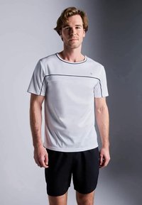 Světle šedé sportovní tričko s černými lemy a krátkým rukávem, spojené s tmavými šortkami. Materiál vypadá prodyšně a hladce.