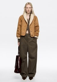 Chaqueta de ante marrón con un cuello de borreguillo color crema, combinada con pantalones de pierna ancha color verde oliva y un bolso de cuero rojo oscuro. Top de punto texturizado debajo.