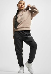 Übergroßer beiger Hoodie mit gesticktem Logo, kombiniert mit dunkelgrauen Jogginghosen mit grafischem Design und schwarzen Kordeln. Weiße Sportschuhe.
