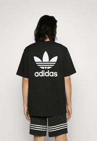 Čierne bavlnené tričko s veľkým bielym logom Adidas a dizajnom trojlístka na chrbte, v kombinácii s očkovanými čiernymi šortkami s bielymi pruhmi.