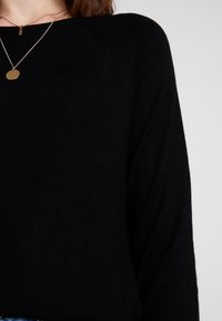 Pull noir en tricot avec une coupe décontractée. Il présente des manches raglan et une texture lisse. Associé à des colliers métalliques superposés.