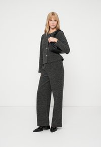 Γκρι πλεκτό κασκόλ με υφή, με κουμπιά snap και αντίστοιχα παντελόνια σε φαρδύ κόψιμο. Το μοντέλο κρατά μια μικρή μαύρη τσάντα και φοράει μυτερές μαύρες γόβες.