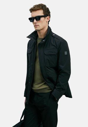 Mann mit schwarzen Sonnenbrillen, dunkler Jacke mit Brusttaschen über einem olivgrünen Pullover, schwarzen Hosen, der eine schwarze Tasche hält und vor einem weißen Hintergrund steht.