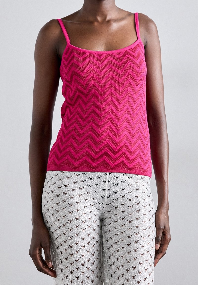 Personne portant un haut sans manches rose à motif zigzag et un pantalon blanc avec un petit motif de cœur noir.