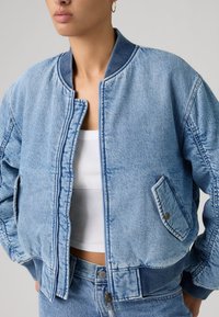 Γυναίκα που φοράει ανοιχτό μπλε τζιν μπουφάν bomber με ριμπ γιακά, μανσέτες και μέση πάνω από λευκό ριμπ αμάνικο τοπ και τζιν που ταιριάζουν.