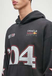 Čierna mikina s kapucňou a predným vreckom, s grafickými prvkami '04' v šedej a červenej farbe a logami Oracle Red Bull Racing. Mäkko sa nosí, vo voľnom strihu.