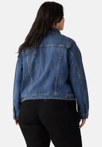 Chaqueta de mezclilla en azul medio, con cuello, cierre de botones y bolsillos frontales. Cortada a la cintura con un ligero desgaste y detalles de costura.