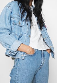 Hellblaues Denim-Jackett mit zwei Brusttaschen, sichtbaren Nähten und Knopfverschluss, kombiniert mit einem weißen T-Shirt und passenden Jeans.