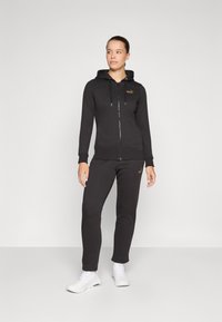 Zwarte zip-up hoodie met gouden accenten, gecombineerd met bijpassende sweatpants. Gemaakt van zachte stof, met een capuchon en geribde manchetten. Witte sneakers.
