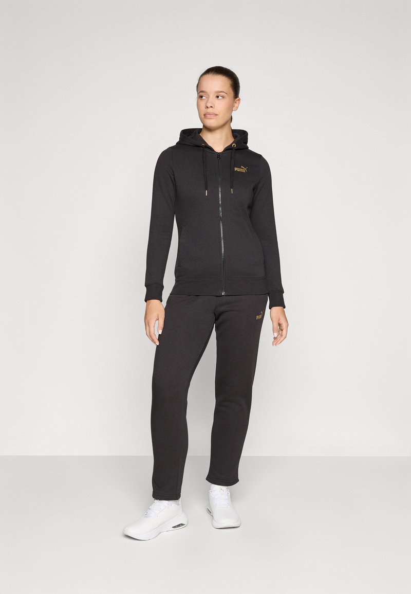 Svart zip-up hoodie med guld-accenter, matchande sweatpants. Tillverkad av mjukt tyg, med huva och ribbad mudd. Vita sneakers.