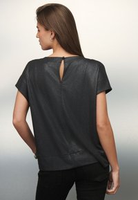 Melnā īsā piedurkņu topā ar maigu spīdumu, ar apaļu kakla izgriezumu un atpakaļ esošu atslēgas caurumu, ko tur ar pogu. Gluda auduma tekstūra.