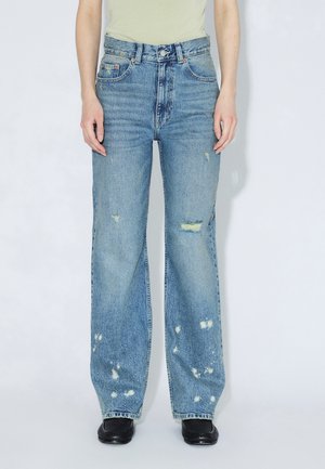 Hellblaue, hoch taillierte Jeans aus Denim mit weitem Bein, Used-Details und sichtbaren Nähten, kombiniert mit schwarzen Schuhen.