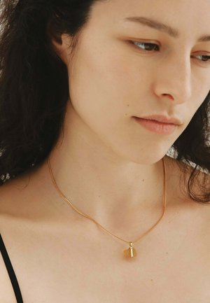 Joven con cabello rizado oscuro que lleva un collar fino de cadena de oro con un colgante de piedra ámbar, mirando hacia abajo y a la derecha.