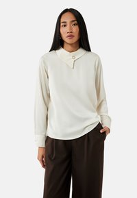 Blusa a maniche lunghe color crema con un design del colletto unico, caratterizzata da un piccolo dettaglio di bottone, abbinata a pantaloni marrone scuro. Morbida al tatto.