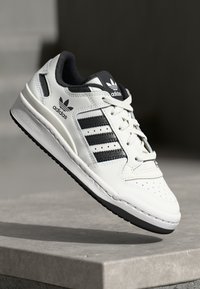 Baskets en cuir blanc avec accents noirs, présentant trois rayures latérales, un bout perforé et une semelle en caoutchouc. Comprend un laçage à l'avant et un logo.