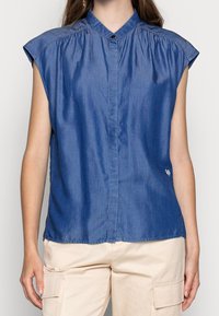 Modrá džínová blůza s krátkým rukávem, mandarínským límcem, zapínáním na knoflíky a nařasením na ramenech; na spodním lemu má malé logo.