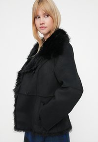 Zwarte jas met een faux fur kraag, twee voorzakken en een knoopsluiting. De textuur lijkt glad met een luxe afwerking bij de kraag.