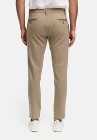 Pantalones elásticos beige de corte ajustado, con dos bolsillos traseros con botones y una textura suave, combinados con zapatillas blancas.