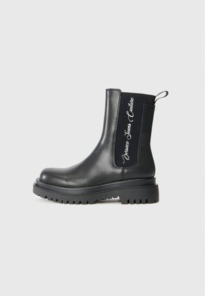 Black leather ankle boots with a chunky sole, featuring elastic side panels and white logo text on one side.