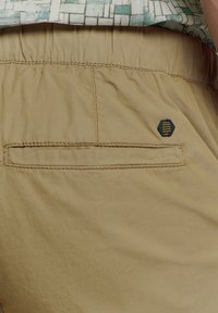 Khaki shorts met een elastische tailleband, voorzien van een achterzak met subtiele stiksels en een zwart hexagonaal embleem detail. Gladde stofstructuur.
