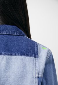 Jeansjacka med tvåfärgsdesign i ljus och mörkblått, med en neongrön accent på axeln. Synliga sömmar.