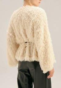 Sweter w kolorze kremowym, o puszystej fakturze, z peplum na dole i dzwonowymi rękawami, z wcięciem w talii, noszony na czarnych skórzanych spodniach.