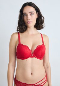 Rode kanten bh met golvende randen, ondersteuning door beugel en verstelbare bandjes. Heeft een decoratieve hanger in het midden.