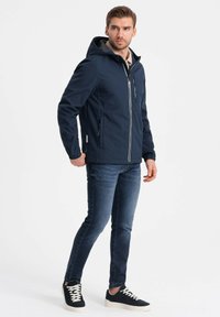 Marineblå hættetrøje med en frontlynlås, sidelommer og grå foring, parret med mørke jeans og sorte sneakers. Enkel og moderne design.