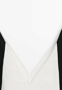 Sort og hvid ærmeløs top med V-hals, der har en struktureret sort stof på skulderen og en glat hvid krop.