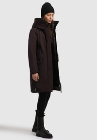 Parka de color marrón oscuro con cuello alto y capucha, presenta grandes bolsillos frontales y un acabado liso mate. Combinada con leggings negros y botas.