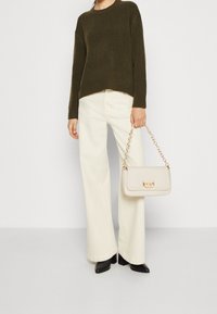 Olivgrön stickad tröja, vita vida byxor, svarta spetsiga skor och en krämfärgad handväska med kedjeförband och rektangulär form.