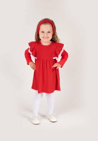 Czerwona sukienka z bufkami z białej koronki, długimi rękawami i marszczonym dołem. Do kompletu białe rajstopy i jasne buty. Dołączona czerwona opaska na głowę.