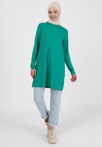 Tunique verte à manches longues avec une coupe décontractée, associée à un jean bleu clair retroussé et des baskets beiges. Design simple avec une texture lisse.