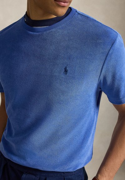 Άντρας φορεί μπλε φωτεινό κοντομάνικο μπλουζάκι από πετσέτα με κεντημένο λογότυπο παίκτη πόλο στο στήθος, από πάνω ένα σκούρο μπλουζάκι με στρογγυλή λαιμόκοψη.