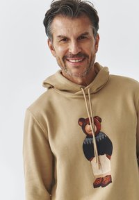 Sudadera beige con un gráfico de un oso en la parte delantera que lleva un suéter negro y pantalones beige. Capucha con cordones y textura de tela suave.