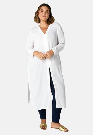 White longline shirt, textured fabric, V-neck, side slits, long sleeves, worn over dark jeans, paired with gold slide sandals.