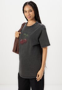 Siva oversized majica s crveno-zelenim grafikom trešnje s točkicama. Model drži smeđi torbu, prikazujući opušteni kroj.