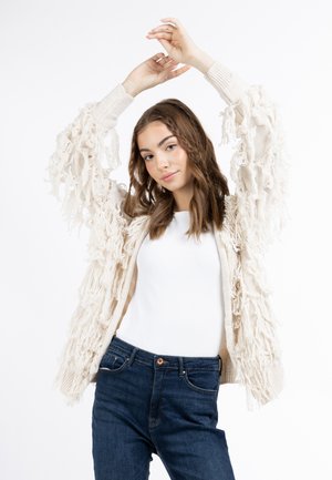 Cardigan crème avec franges en tissu texturé et superposé. Style tricot épais, ouverture sur le devant et poignets côtelés ; porté sur un simple top blanc et un jean foncé.