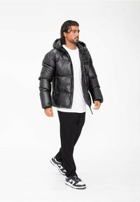 Doudoune noire avec une finition brillante, capuche et coutures bien visibles. Portée sur une chemise blanche, associée à un pantalon noir et des baskets noir et blanc.